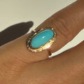Gold ring with a turquoise stone on a finger against a neutral background