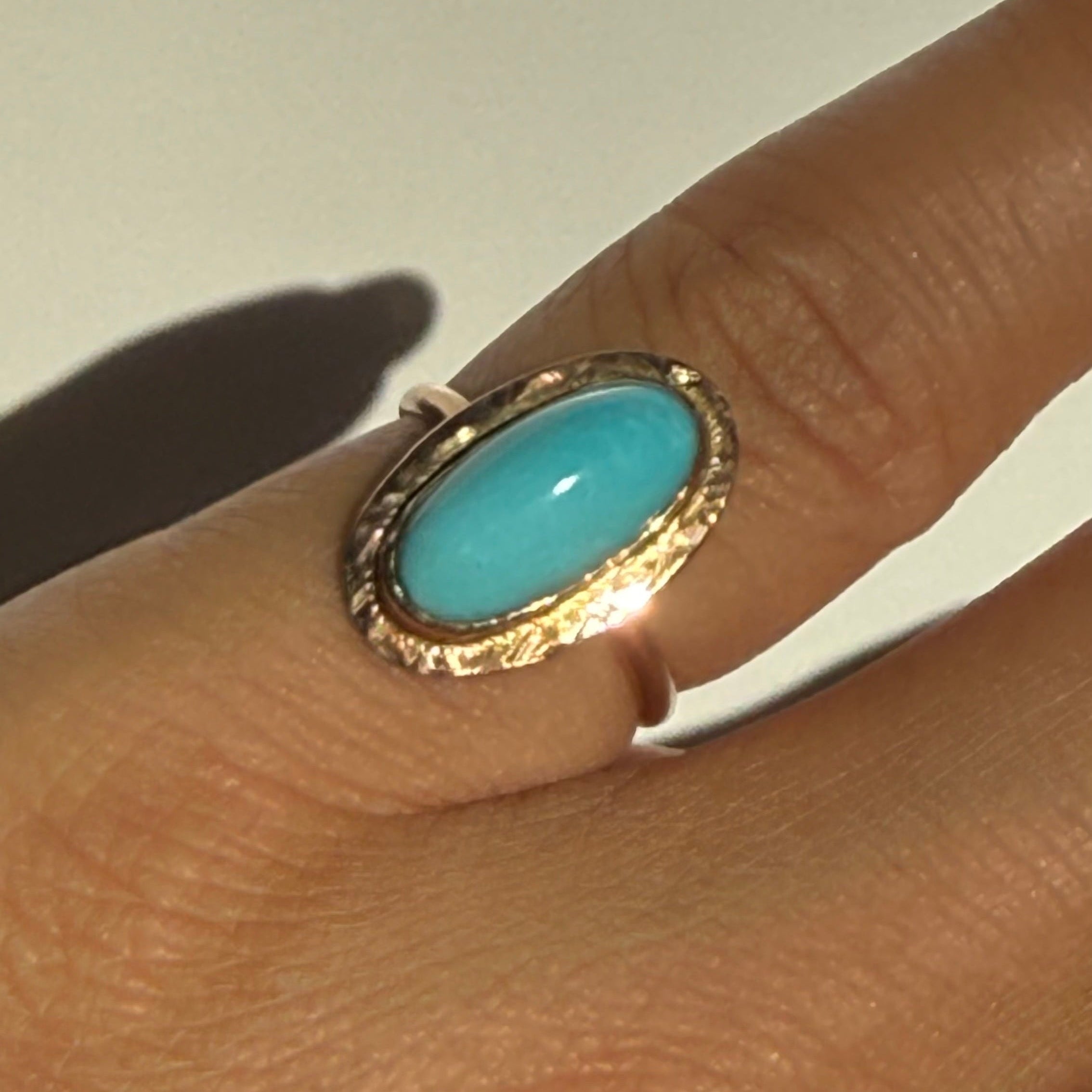 Gold ring with a turquoise stone on a finger against a neutral background
